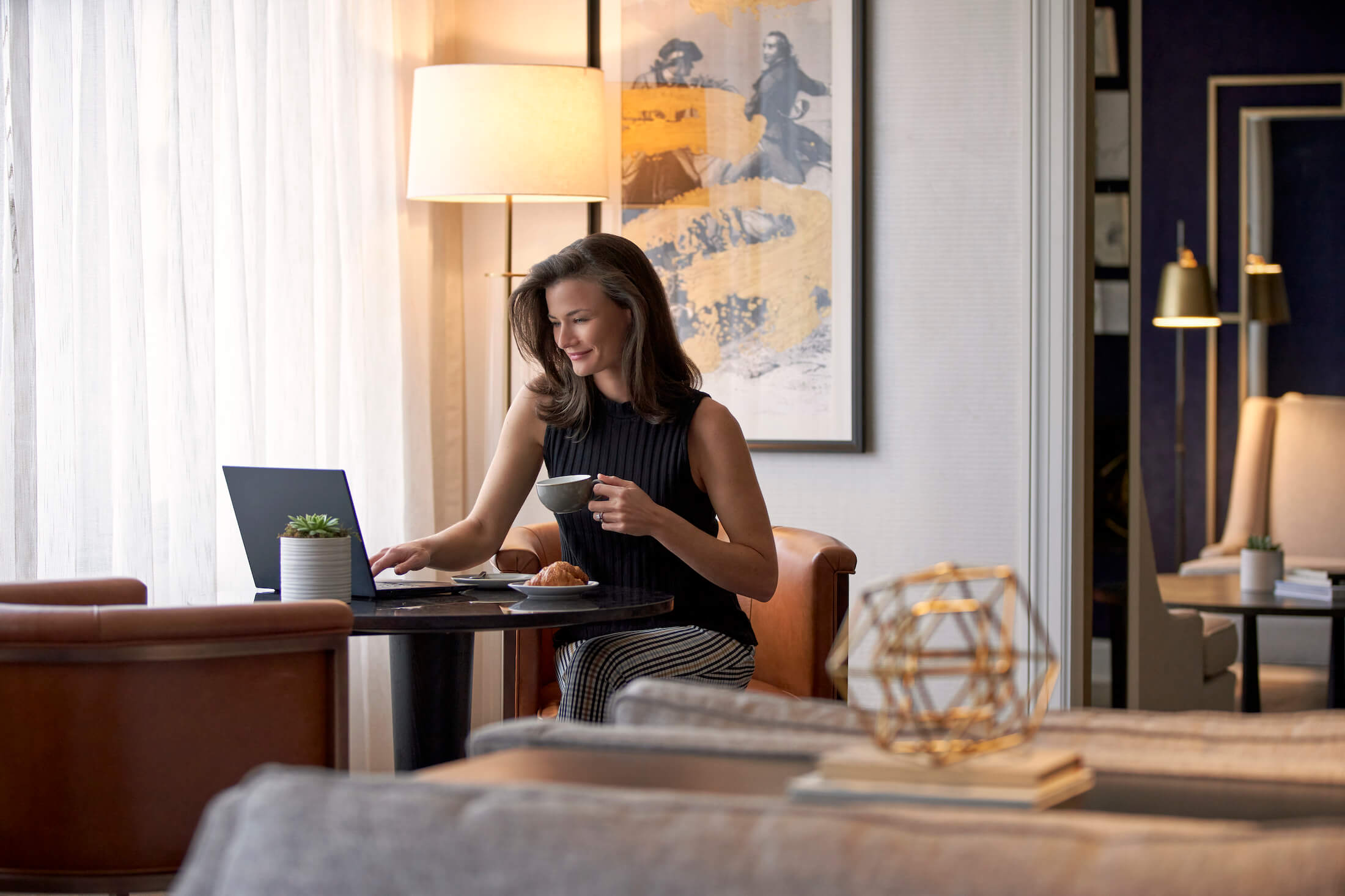 A woman in a hotel room drinking coffee while on a laptop.