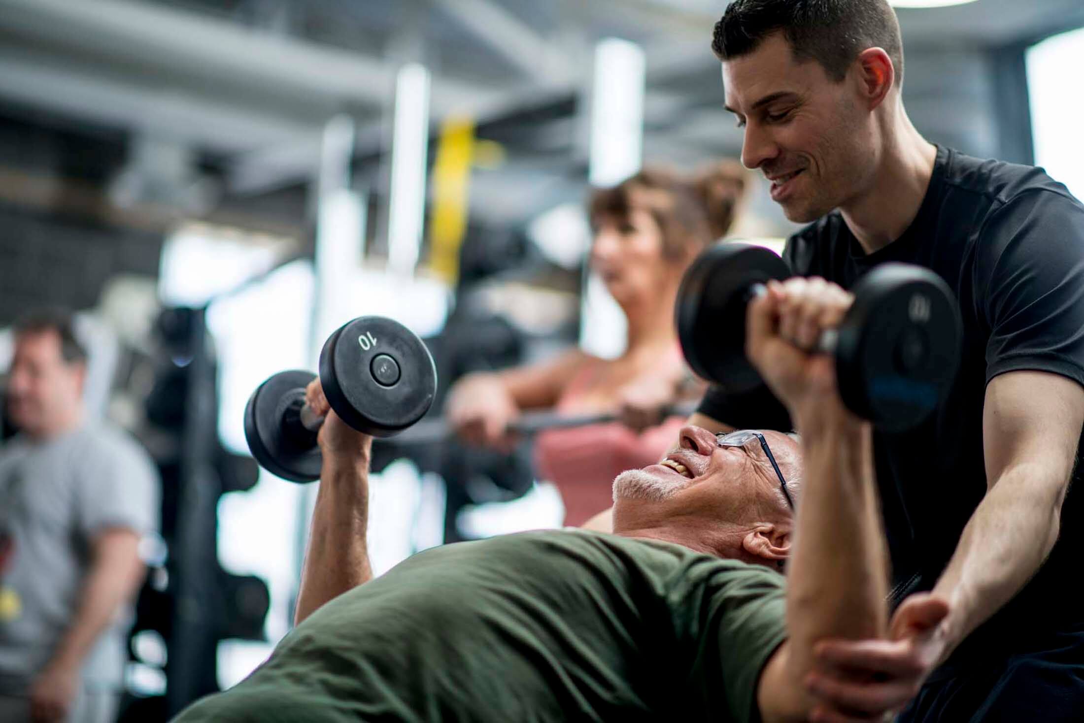 A personal training spotting an elderly man doing dumbbell chess presses at a gym.