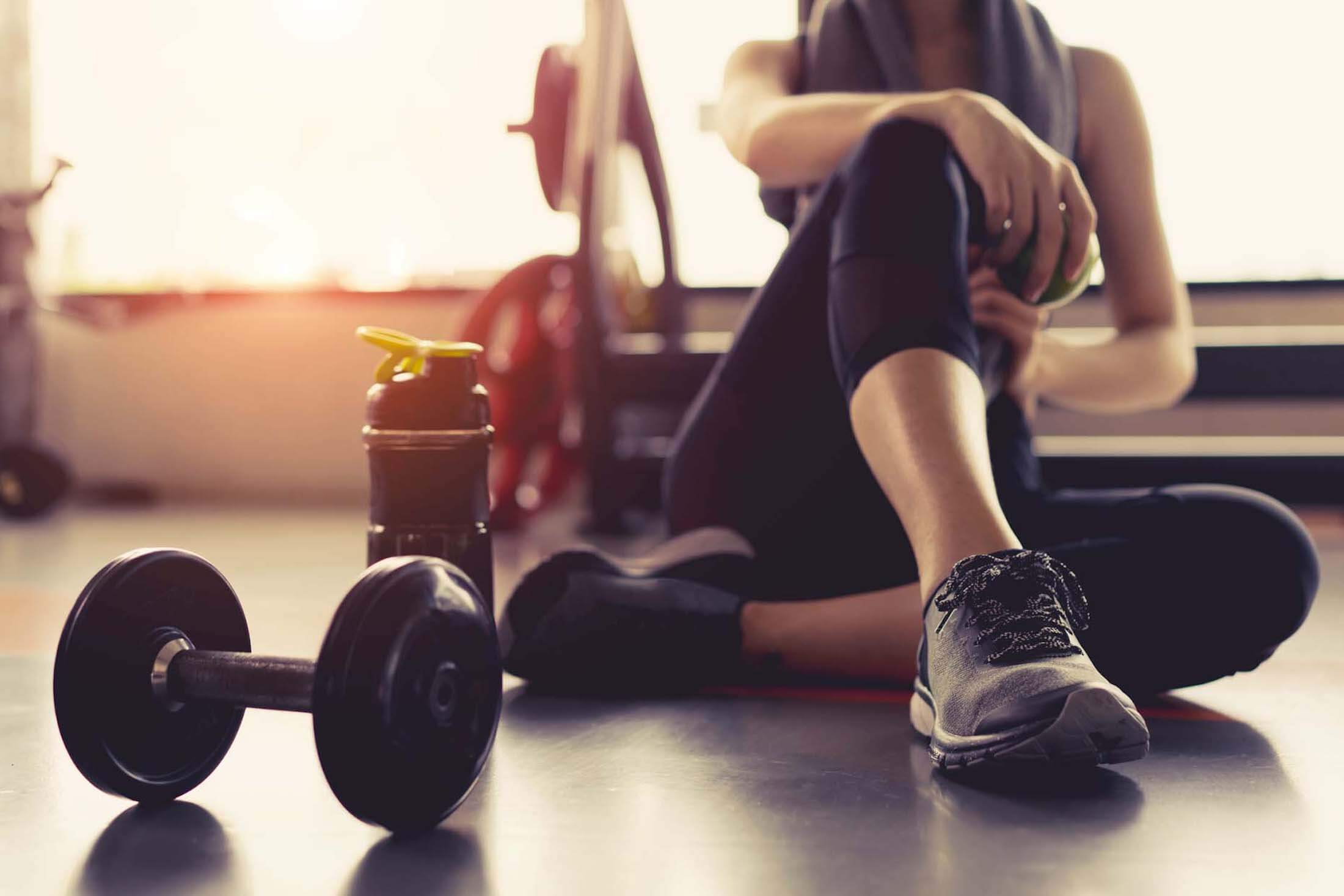 A person sitting on the floor of a gym near a water bottle and a dumbbell.